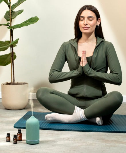 Woman meditating on yoga mat with essential oil diffuser releasing mist – home aromatherapy for relaxation and stress relief