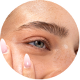 A close-up of a person's eye and hand, showing a blue-gray eye with a defined eyebrow and manicured fingernails, set against a solid black background.