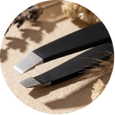 A close-up of a pair of black-handled grooming tools, likely facial or body razors, resting on a textured beige surface with dried botanical elements.
