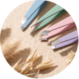 A circular close-up of pastel-colored pencils arranged on a sandy surface with small dried grasses in the foreground.
