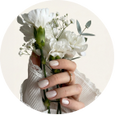 A close-up of a hand holding a bouquet of white flowers with green stems and small white blossoms, framed inside a light beige circular border.