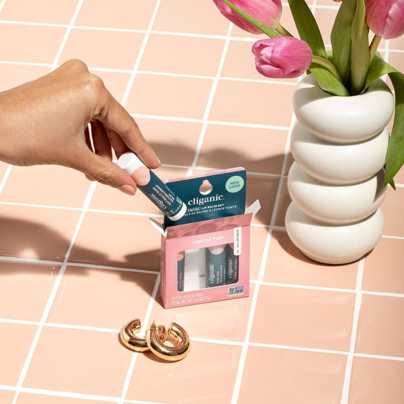 Hand holding Cliganic lip balm gift set in pink packaging on tiled surface with pink tulips and gold earrings.